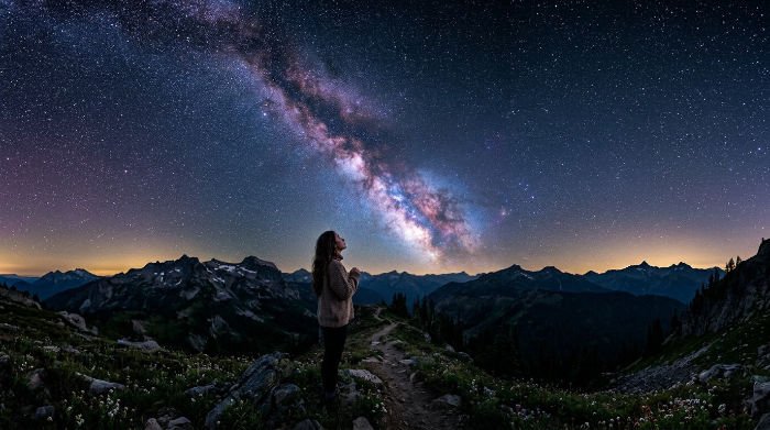 Small figure standing on a mountain ridge at night, gazing up at the Milky Way arching across the sky — when you speak to the universe, who listens