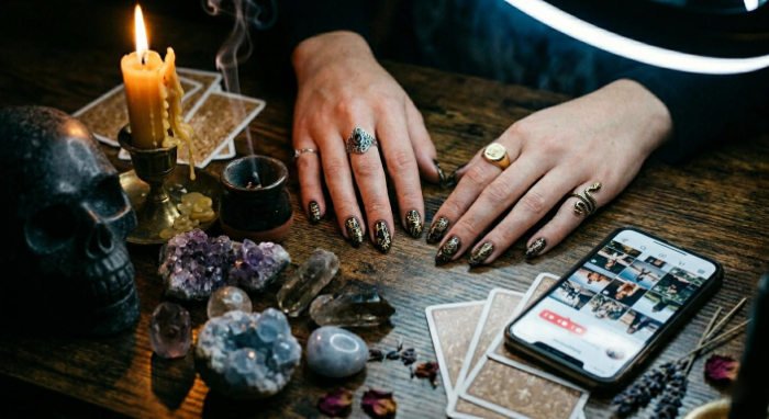 Hands with black nail polish on a table with candles, skull, crystals, tarot cards and an iPhone with Instagram open