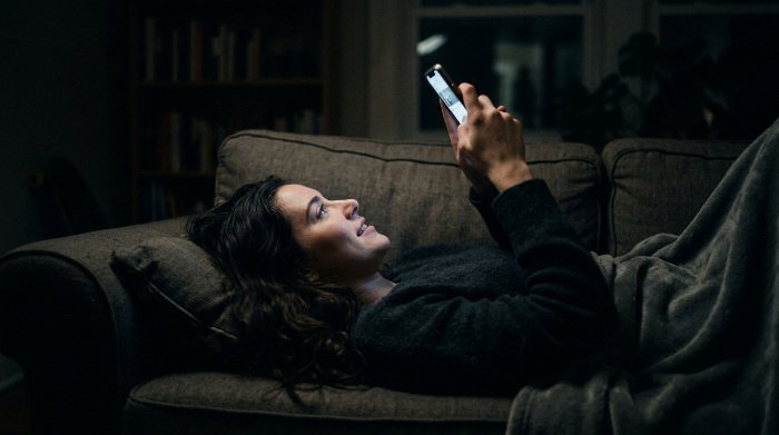 Woman lying on a sofa at night looking at her phone, illuminated by the screen, in the moment of the Wow Effect